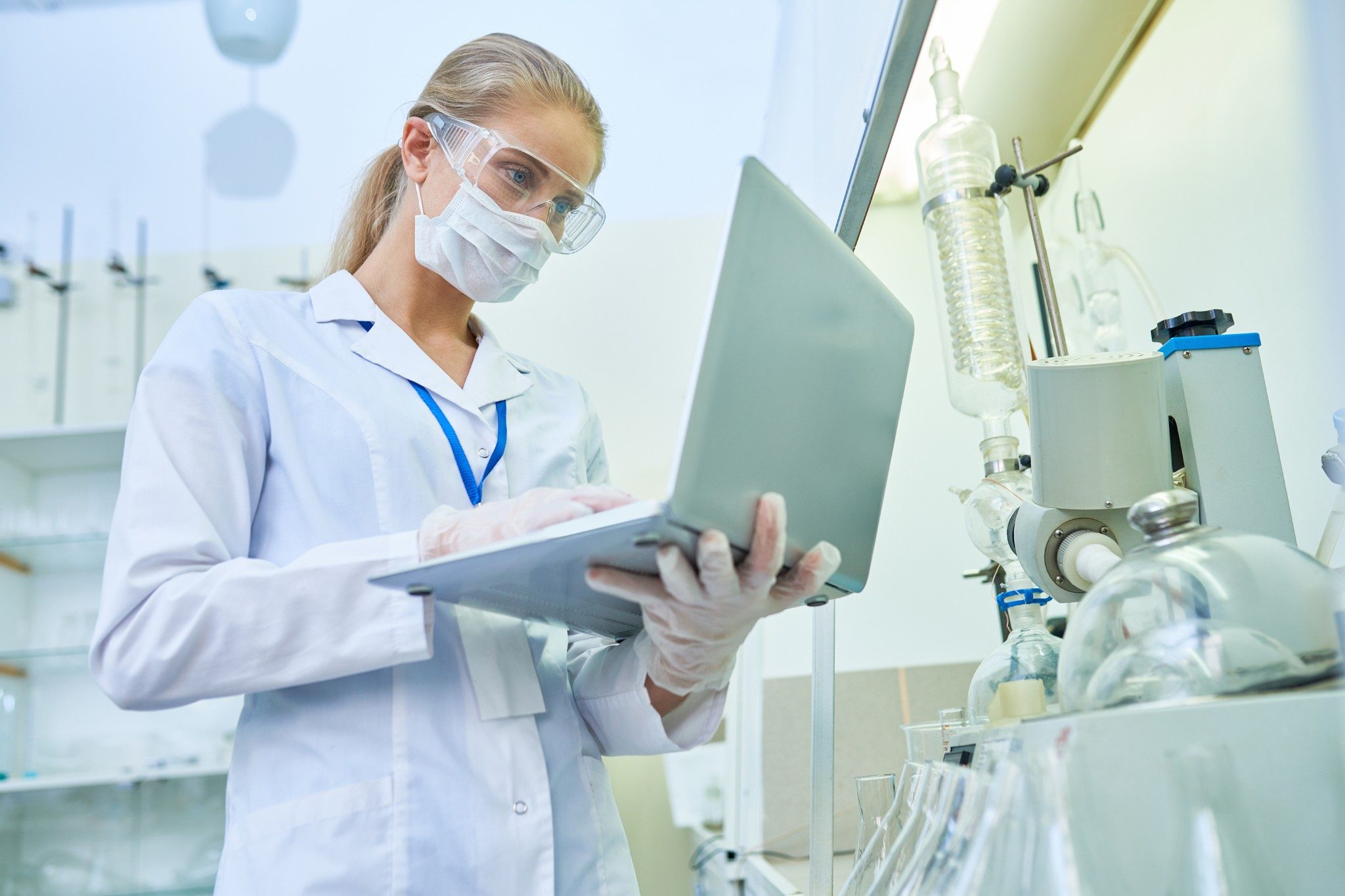 Una investigadora joven con rostro serio y confianza, con máscara de protección y gafas de seguridad, utilizando un equipo portátil para examinar el archivo con el resultado del experimento mientras trabaja con el equipo médico